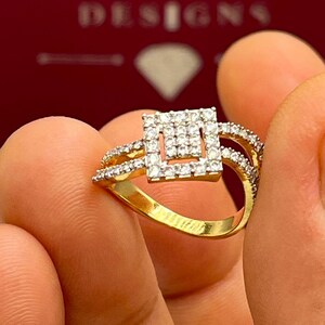 Puede incluir: Un anillo de oro con un dise&ntilde;o cuadrado central incrustado con peque&ntilde;as piedras preciosas claras. El anillo tiene una banda dividida con piedras preciosas adicionales. El anillo se sostiene entre los dedos, con la marca "HANSA DESIGNS" visible en el fondo.
