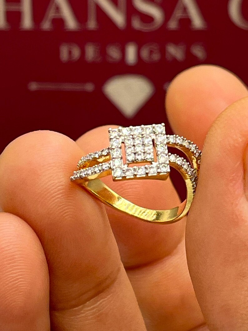 Puede incluir: Un anillo de oro con un dise&ntilde;o cuadrado central incrustado con peque&ntilde;as piedras preciosas claras. El anillo tiene una banda dividida con piedras preciosas adicionales. El anillo se sostiene entre los dedos, con la marca "HANSA DESIGNS" visible en el fondo.