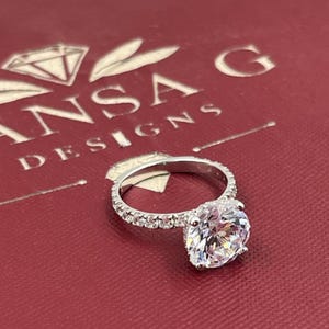 May include: A silver ring with a large, pear-shaped, clear gemstone. The band is encrusted with smaller, clear gemstones. The ring sits on a red surface with the text "ANSA DESIGNS".