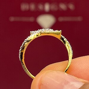 Puede incluir: Primer plano de un anillo de oro con un grupo de diamantes cuadrados y diamantes m&aacute;s peque&ntilde;os a lo largo de la banda. El anillo se sostiene sobre un fondo rojo, mostrando los detalles de la joya.