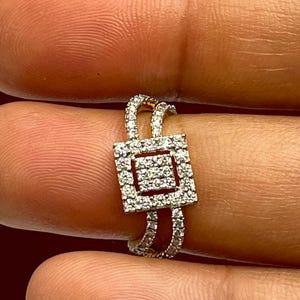 Puede incluir: Un anillo de oro con un centro cuadrado con incrustaciones de diamantes. El anillo presenta un dise&ntilde;o de doble banda, con diamantes m&aacute;s peque&ntilde;os engastados a lo largo de las bandas. El anillo se sostiene entre dos dedos.