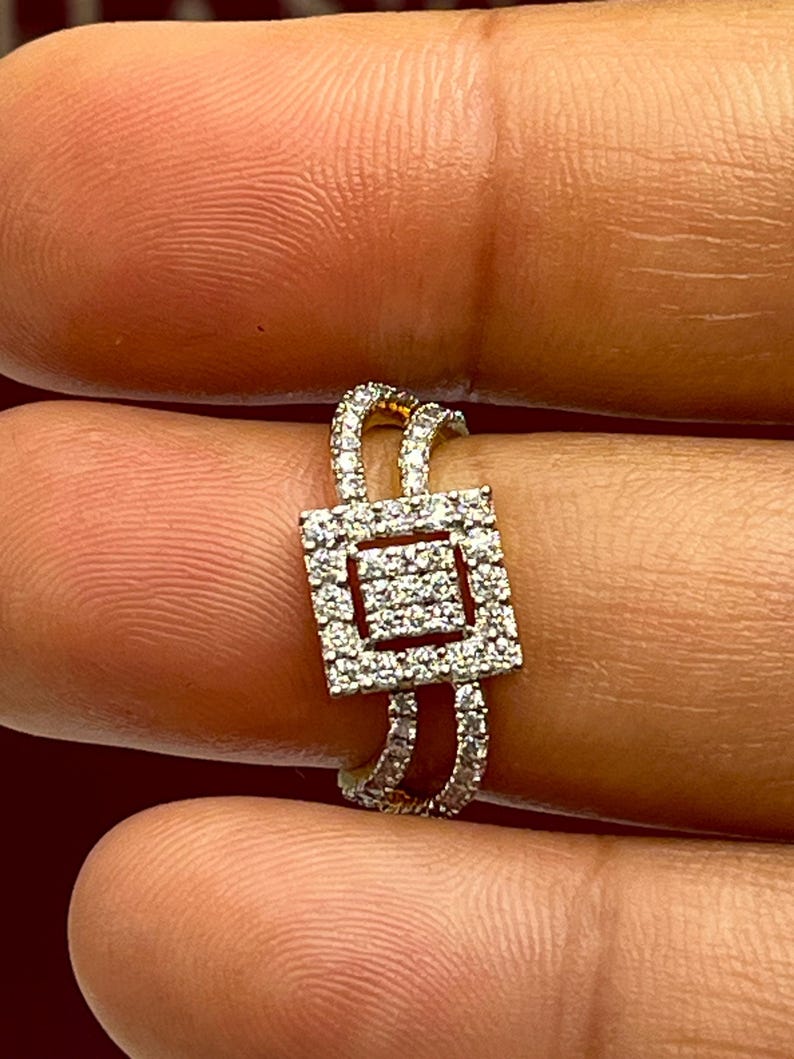 Puede incluir: Un anillo de oro con un centro cuadrado con incrustaciones de diamantes. El anillo presenta un dise&ntilde;o de doble banda, con diamantes m&aacute;s peque&ntilde;os engastados a lo largo de las bandas. El anillo se sostiene entre dos dedos.