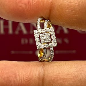 Puede incluir: Un anillo dorado con un centro cuadrado con diamantes y diamantes m&aacute;s peque&ntilde;os a lo largo de la banda. El anillo se sostiene entre los dedos, mostrando su intrincado dise&ntilde;o y brillo. El dise&ntilde;o del anillo es geom&eacute;trico y elegante.