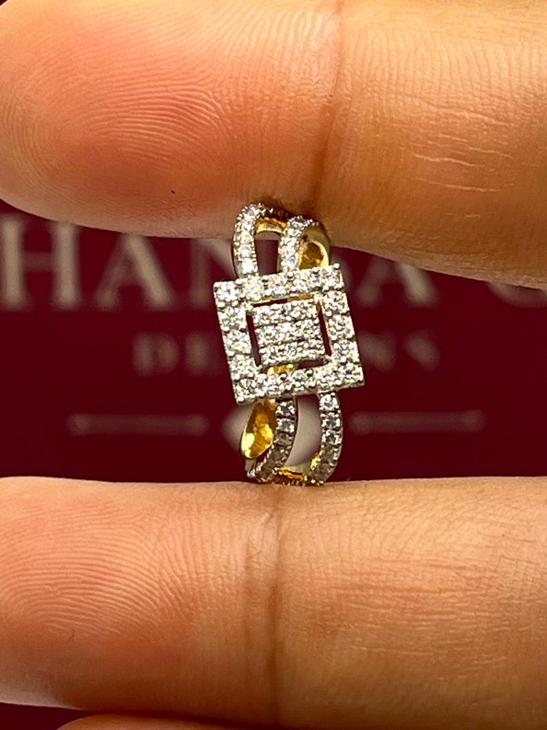 Puede incluir: Un anillo dorado con un centro cuadrado con diamantes y diamantes m&aacute;s peque&ntilde;os a lo largo de la banda. El anillo se sostiene entre los dedos, mostrando su intrincado dise&ntilde;o y brillo. El dise&ntilde;o del anillo es geom&eacute;trico y elegante.