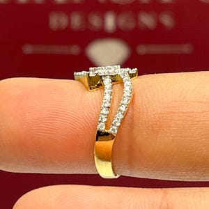 Puede incluir: Un anillo de oro con una parte superior cuadrada con diamantes y una banda dividida. El anillo presenta una doble fila de peque&ntilde;os diamantes a lo largo de la banda, creando un efecto brillante. El anillo se muestra sobre un fondo rojo.