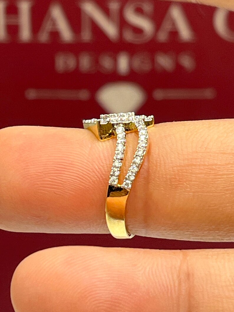 Puede incluir: Un anillo de oro con una parte superior cuadrada con diamantes y una banda dividida. El anillo presenta una doble fila de peque&ntilde;os diamantes a lo largo de la banda, creando un efecto brillante. El anillo se muestra sobre un fondo rojo.