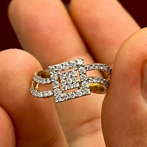 Puede incluir: Un anillo de oro con un grupo cuadrado de diamantes en el centro, rodeado de diamantes m&aacute;s peque&ntilde;os. El anillo tiene un dise&ntilde;o &uacute;nico con detalles calados y se sostiene entre los dedos.