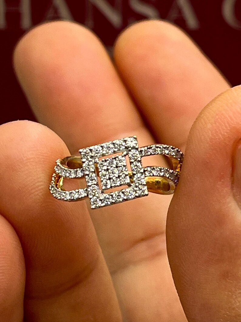 Puede incluir: Un anillo de oro con un grupo cuadrado de diamantes en el centro, rodeado de diamantes m&aacute;s peque&ntilde;os. El anillo tiene un dise&ntilde;o &uacute;nico con detalles calados y se sostiene entre los dedos.