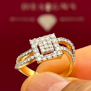 Puede incluir: Un anillo de oro con un centro de diamantes cuadrados y diamantes m&aacute;s peque&ntilde;os a lo largo de la banda. El anillo se sostiene sobre un fondo rojo con el texto "HANSA DESIGNS".