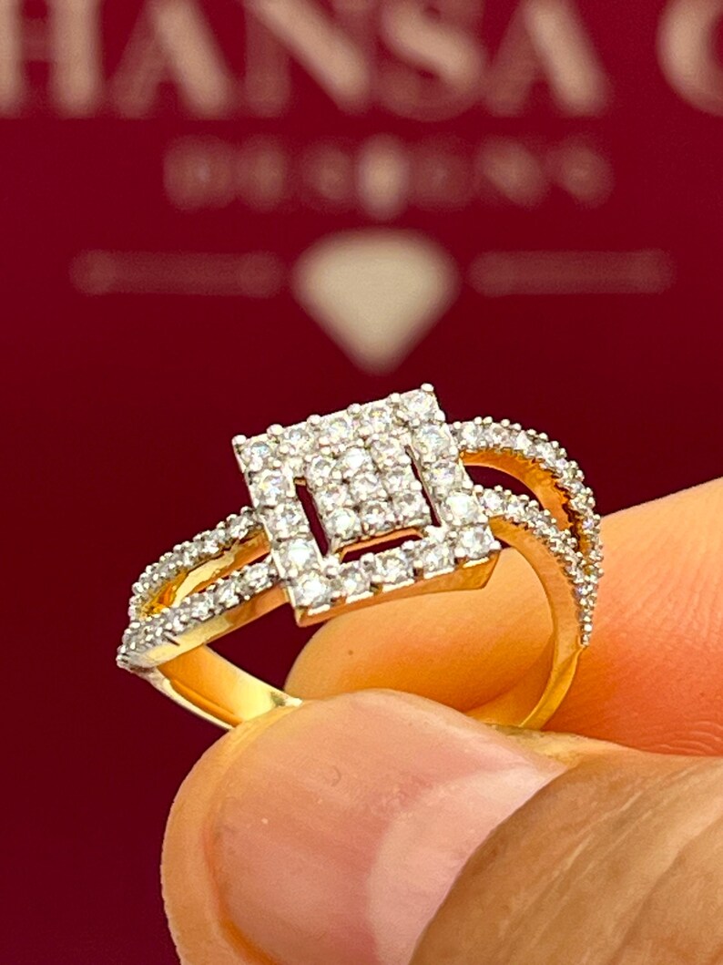 Puede incluir: Un anillo de oro con un centro de diamantes cuadrados y diamantes m&aacute;s peque&ntilde;os a lo largo de la banda. El anillo se sostiene sobre un fondo rojo con el texto "HANSA DESIGNS".