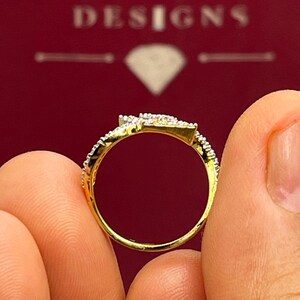Puede incluir: Un anillo de oro con un dise&ntilde;o en forma de diamante, incrustado con peque&ntilde;as piedras brillantes. El anillo se sostiene entre dos dedos, mostrando sus intrincados detalles sobre un fondo rojo. El nombre de la marca "HANSA DESIGNS" es visible.