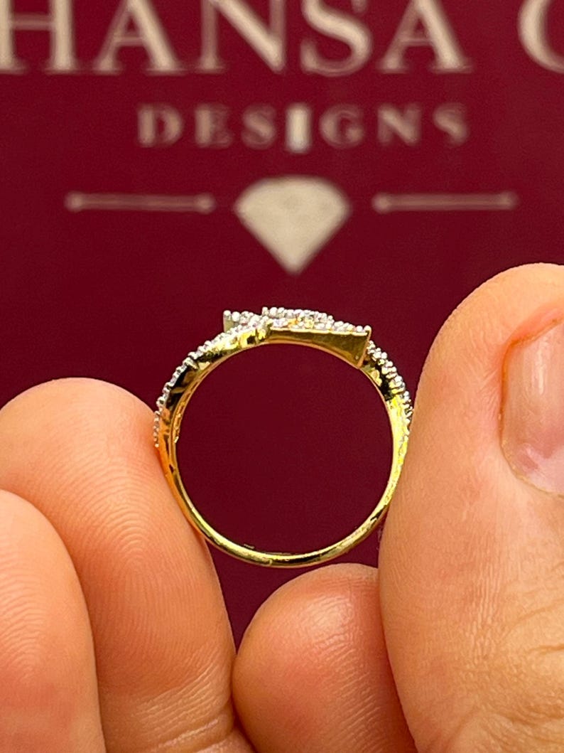 Puede incluir: Un anillo de oro con un dise&ntilde;o en forma de diamante, incrustado con peque&ntilde;as piedras brillantes. El anillo se sostiene entre dos dedos, mostrando sus intrincados detalles sobre un fondo rojo. El nombre de la marca "HANSA DESIGNS" es visible.
