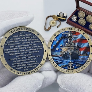 Op de afbeelding: Twee bronzen herdenkingsmunten met tekst en afbeeldingen die betrekking hebben op de Amerikaanse marine. Eén munt toont de tekst "Sailor's Creed" en de andere toont een schip met een Amerikaanse vlag. Extra items met marinethema zijn zichtbaar.