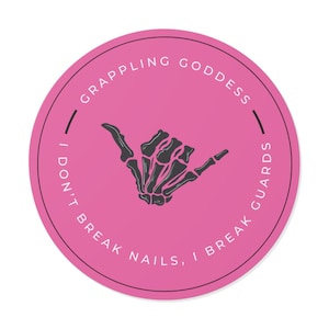 Könnte beinhalten: Ein runder, pinkfarbener Aufkleber mit dem Text "GRAPPLING GODDESS" und "I DON'T BREAK NAILS, I BREAK GUARDS". In der Mitte befindet sich eine schwarz-graue Skeletthand im Shaka-Zeichen.