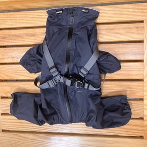 Op de afbeelding: Een marineblauwe hondenregenjas met een ontwerp over het hele lichaam. De jas heeft een capuchon, een lange ritssluiting en grijze verstelbare bandjes met metalen gespen. De poten hebben elastische manchetten. De regenjas beschermt honden tegen regen.