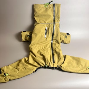 May include: Mustard-colored dog raincoat with a hood, black zipper, and neon green accents. The dog coat has sleeves for the front legs and a drawstring at the hood. The coat is made of a waterproof material.