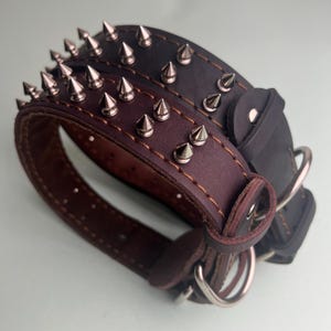 Puede incluir: Se muestran dos collares de cuero para perros, uno marrón oscuro y otro negro. Cada collar está adornado con filas de púas metálicas de color plateado. Los collares tienen anillas en D metálicas y están cosidos con hilo en contraste.