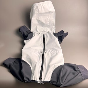 Peut inclure: Un imperméable pour chien blanc et gris avec capuche. La veste a une fermeture éclair noire sur toute la longueur et des détails noirs sur les manches et les jambes. L'imperméable est conçu pour couvrir le corps et les pattes du chien.