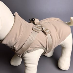 Puede incluir: Un abrigo para perros de color beige con arnés integrado. El abrigo es acolchado y tiene un cuello alto. El arnés tiene correas ajustables y hebillas de plástico. El abrigo para perros está diseñado para mantener a los perros abrigados y cómodos.