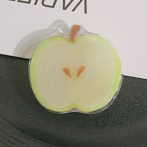 May include: A clear, apple-shaped sticker with a green outline and a light yellow center, featuring a brown stem and two brown seeds. The sticker is placed on a black plate, with a white card behind it that reads "VARIETY IS THE SPICE OF LIFE."