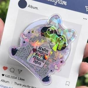 May include: A clear, iridescent raccoon-shaped phone grip with the text "ALWAYS FREAKIN BUSY READING TRASH" on a book. The grip is on a blue Instagram-themed card with a heart and like icons.