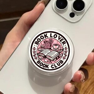 May include: A white smartphone with a pop-up grip featuring a pink and white design. The grip reads "BOOK LOVER BOOK CLUB" around an illustration of an open book, a cup, and flowers. The phone is held over a wooden surface.