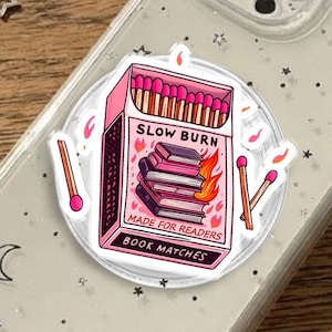 May include: A clear phone case with a decorative sticker featuring a pink matchbox design. The matchbox reads "SLOW BURN" and "MADE FOR READERS BOOK MATCHES". A silver star necklace is in the background.