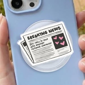 May include: A light blue phone case with a white pop socket featuring a newspaper design. The newspaper graphic reads "BREAKING NEWS" with the headline "Girl falls in love with another fictional character."