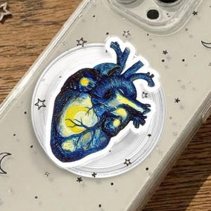 May include: Clear phone case with a blue and yellow anatomical heart design. The case has a glittery finish with star and crescent moon accents. A silver necklace with a star pendant is also visible.