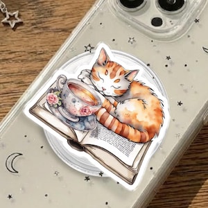 May include: Clear phone case with a whimsical design featuring a sleeping orange tabby cat curled up on a book next to a teacup. The case has a glittery, starry pattern with crescent moons and stars.