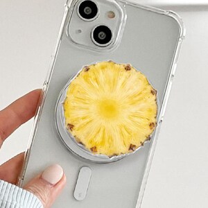 May include: A clear phone case with a yellow pineapple slice phone grip. The phone case has a dual-lens camera and a white grip base. The pineapple slice is yellow with a brown rind.