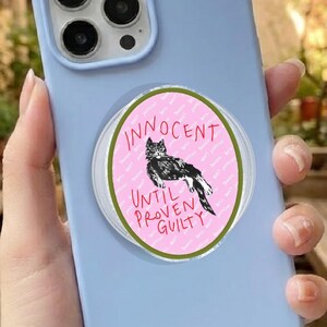 May include: A pale blue phone case with a pop socket. The pop socket has a pink background, a black and white cat illustration, and the text "Innocent Until Proven Guilty" in a green circular design.