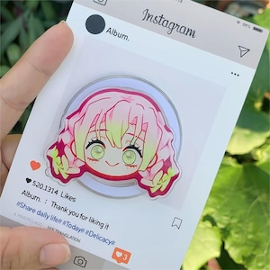 May include: A clear acrylic phone grip with a pink and green anime character design. The grip is circular and set against a white background with an Instagram-style frame. The text reads "Album. Thank you for liking it".