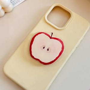 May include: A light yellow phone case with a clear, apple-shaped pop socket. The pop socket features a red outline and a pink interior with two small, dark brown seeds. The phone case has a smooth finish.
