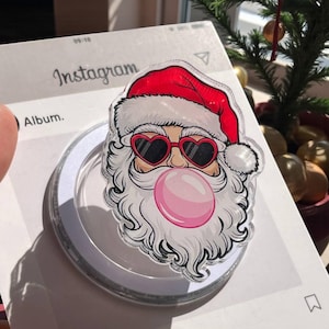 May include: A clear phone grip featuring a Santa Claus design with heart-shaped sunglasses and a pink bubble gum bubble. The Santa wears a red hat and has a white beard. The grip is on a white surface with the Instagram logo visible.