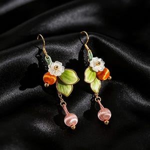 May include: A pair of drop earrings featuring gold-tone hooks, white flower accents, green leaves, orange spherical beads, and light pink dangling beads. The design incorporates delicate thread-wrapped elements and small gold-tone metal beads.