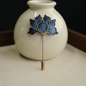 May include: A navy blue floral lapel pin with gold-colored accents. The pin features a detailed flower design with a long, straight pin and a decorative end. The pin is displayed against a cream-colored background.