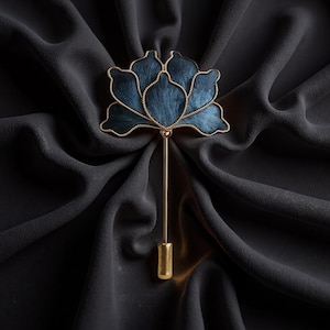 May include: A blue flower-shaped lapel pin with gold-coloured trim and a long, straight pin. The flower has a textured appearance and is set against a dark, draped fabric background. The pin is a stylish accessory.