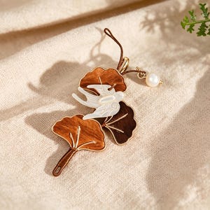 Op de afbeelding: Een decoratieve hanger met twee bruine ginkgobladvormen met goudkleurige omlijningen, een witte iriserende vogelvorm die er bovenop ligt, en een kleine witte parel bevestigd aan een dunne bruine koordlus voor ophanging of bevestiging.