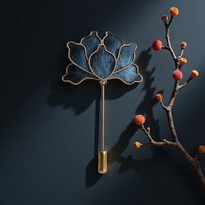 May include: A decorative wall hanging in the shape of a blue lotus flower with gold trim and a long gold stem. The flower is mounted on a dark blue wall, with a branch of orange and yellow buds to the right.