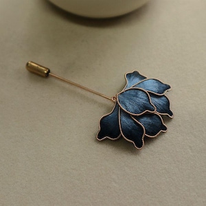 May include: A floral lapel pin with a dark blue flower design and a gold-colored pin. The flower has a textured appearance with gold-colored trim. The pin is a stylish accessory.