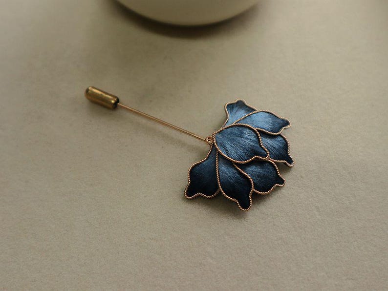 May include: A floral lapel pin with a dark blue flower design and a gold-colored pin. The flower has a textured appearance with gold-colored trim. The pin is a stylish accessory.