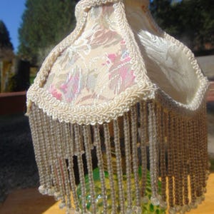 May include: A lamp shade featuring a floral fabric pattern in shades of pink, green, and cream, finished with a cream-colored braided trim and a long fringe of clear, translucent beads hanging from the bottom edge of the square-shaped shade.
