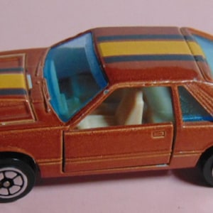 May include: A die-cast model car in a reddish-brown color with yellow and black stripes on the hood and roof. The car has black wheels with white accents and clear blue windows. The interior is a light beige color.