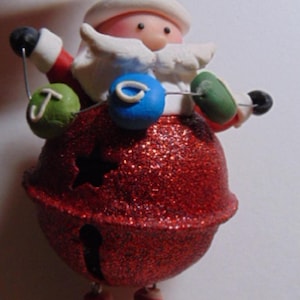 May include: A Christmas ornament shaped like Santa Claus holding a string of colorful ornaments. Santa wears a red hat and coat, with a glittery red bell-shaped body. The ornament has striped legs and black boots.