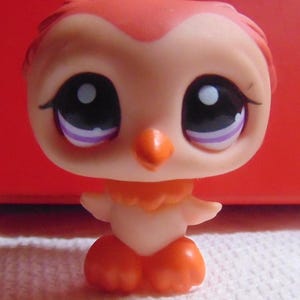 May include: A small, orange and peach-colored owl figurine with large, expressive eyes. The owl has a round body, small wings, and orange feet. The eyes are black with white highlights and purple accents. The figurine is set against a red background.