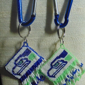 May include: Two blue carabiner keychains with white and green beaded keychains featuring a blue and white logo of a bird with a green eye.