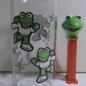 May include: A clear glass tumbler with cartoon frog graphics and snowflakes. Next to it is a green frog PEZ dispenser with an orange base. The tumbler has two green frog characters wearing white shirts and sneakers.