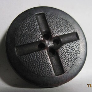 May include: A black plastic button with a cross design and four holes. The button has a textured surface.