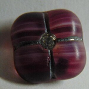May include: A purple and white glass button with a clear rhinestone in the center. The button has a four-pointed star shape.